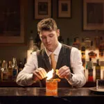 Man lighting a drink at 12.18. Roxburghe Hotel's historical Bar 1745, symbolizing tradition and elegance.