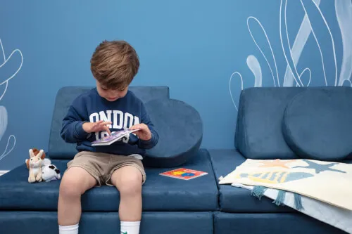 Young boy reading a book on a blue sofa with toys and a decorative blanket.