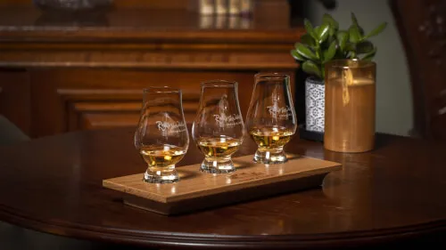 Three whiskey glasses on a wooden tray, accompanied by a plant and candle in a cozy setting.