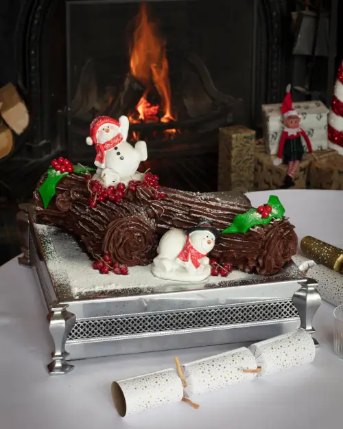 Festive chocolate Yule log cake with snowmen, holly, and a cozy fireplace background.